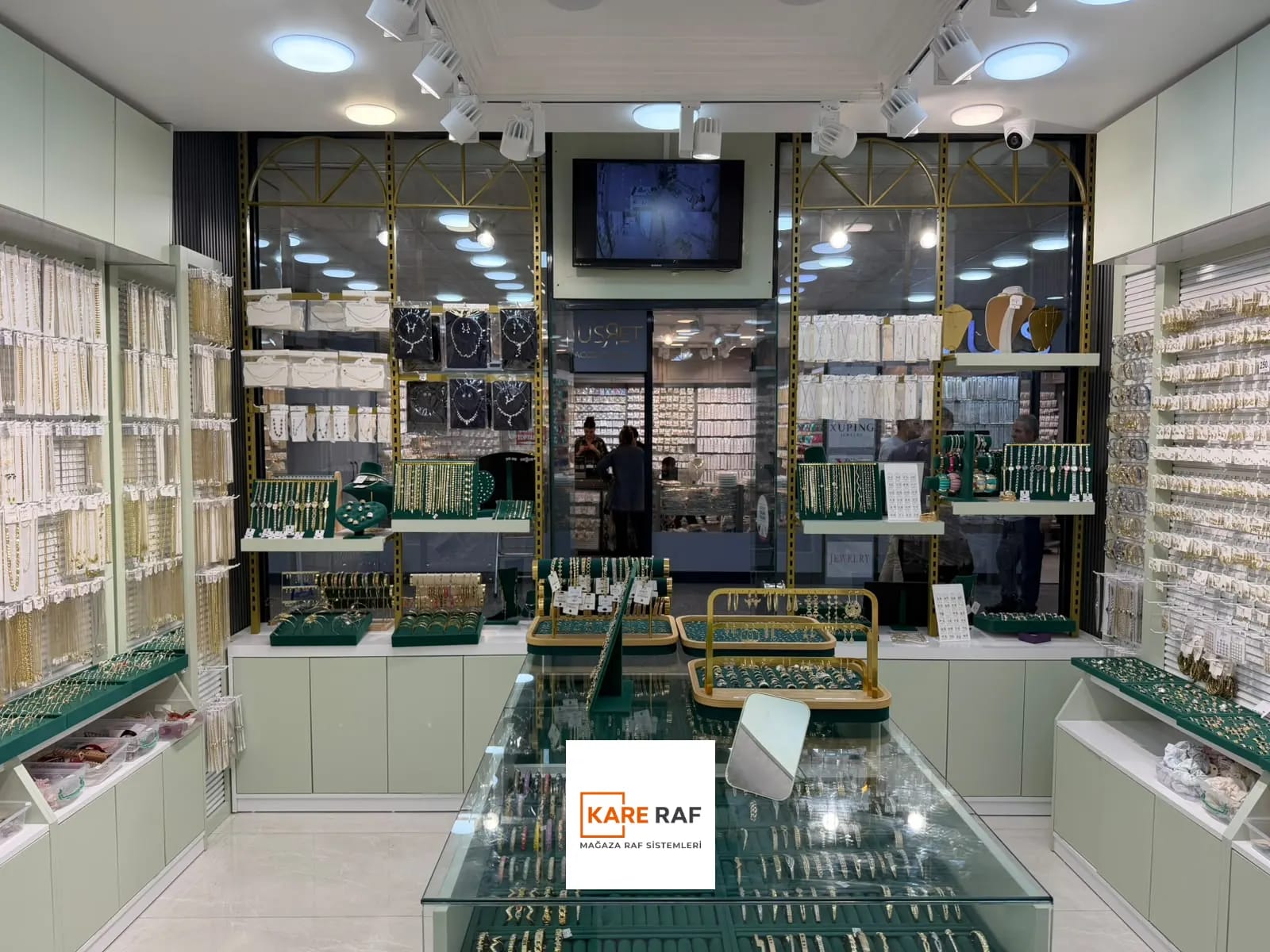 İçeriden vitrine bakış: cam üstlü teşhir tezgâhı, kanallı pano duvarlar ve adaçayı yeşili alt dolaplar